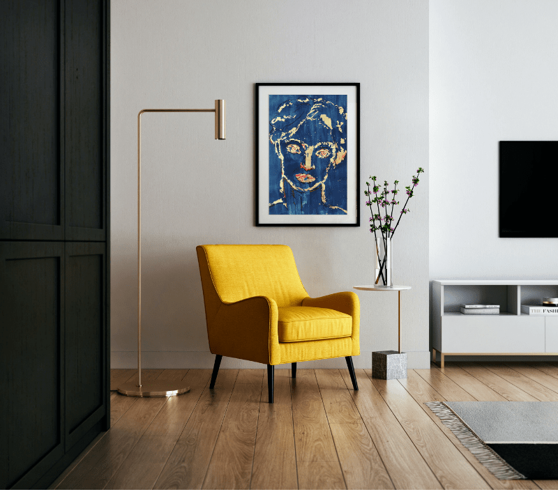 A simple, elegant modern interior with wide oak plank flooring and soft grey walls. On the wall is hung an original painting by Louisa Currier showing a face picked out in gold leaf against a deep blue background. Below the painting is a yellow chair, a sleek brass lamp alongside.